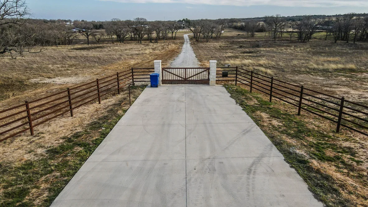 Concrete driveway with metal ranch gate and pipe fencing at rural property entrance