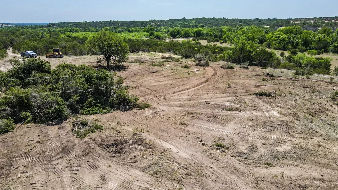 Graded dirt site with cleared pathways, heavy equipment, and selective tree retention