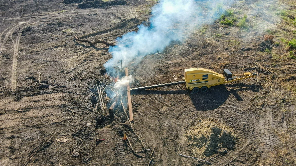 Yellow wood chipper processing tree debris on cleared land with smoke visible from active chipping operation