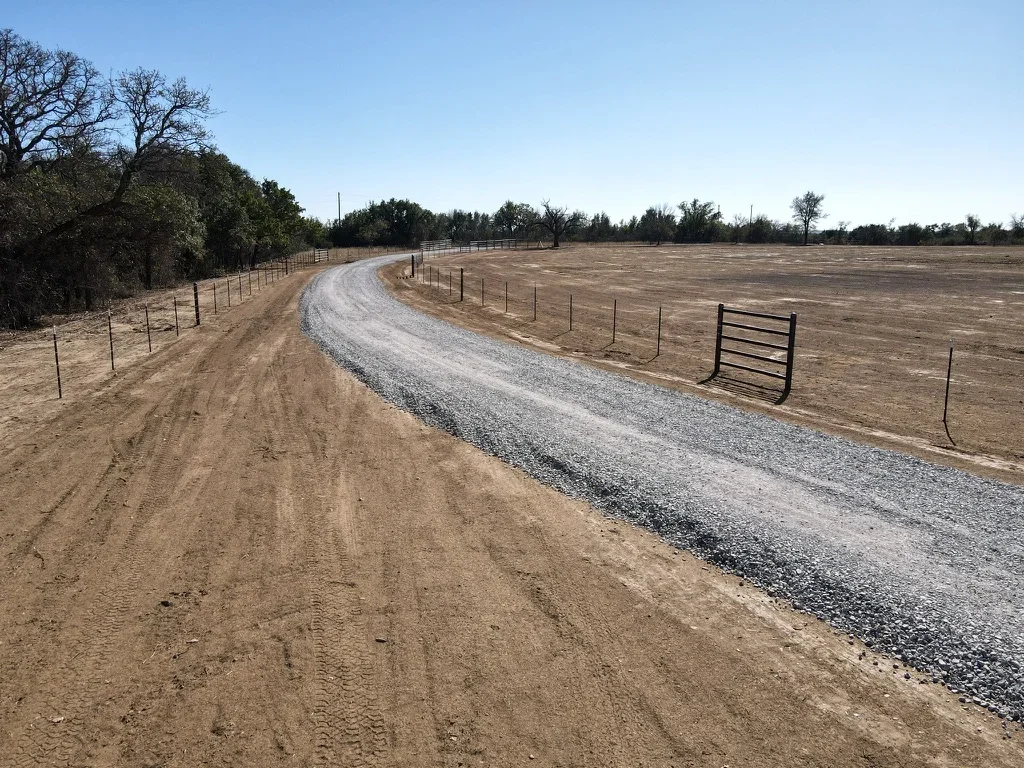 Curved gravel driveway with compacted soil base and crushed stone surface, bordered by post-and-rail fencing