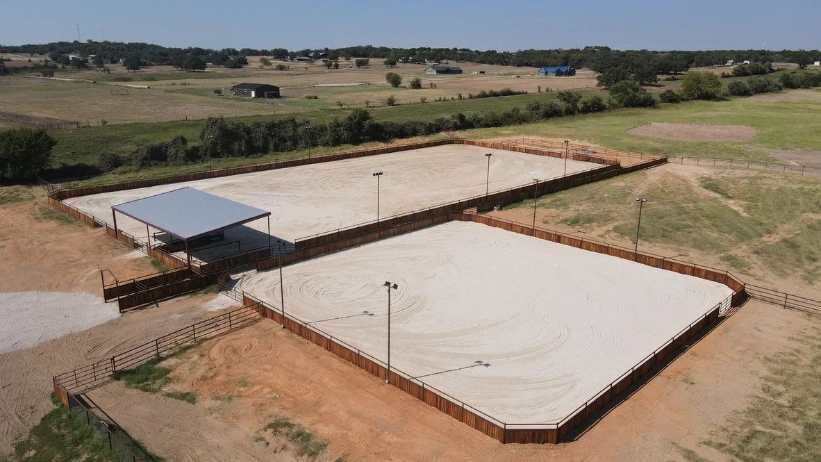 Two sand arenas with metal fencing and lighting poles on prepared earthwork site