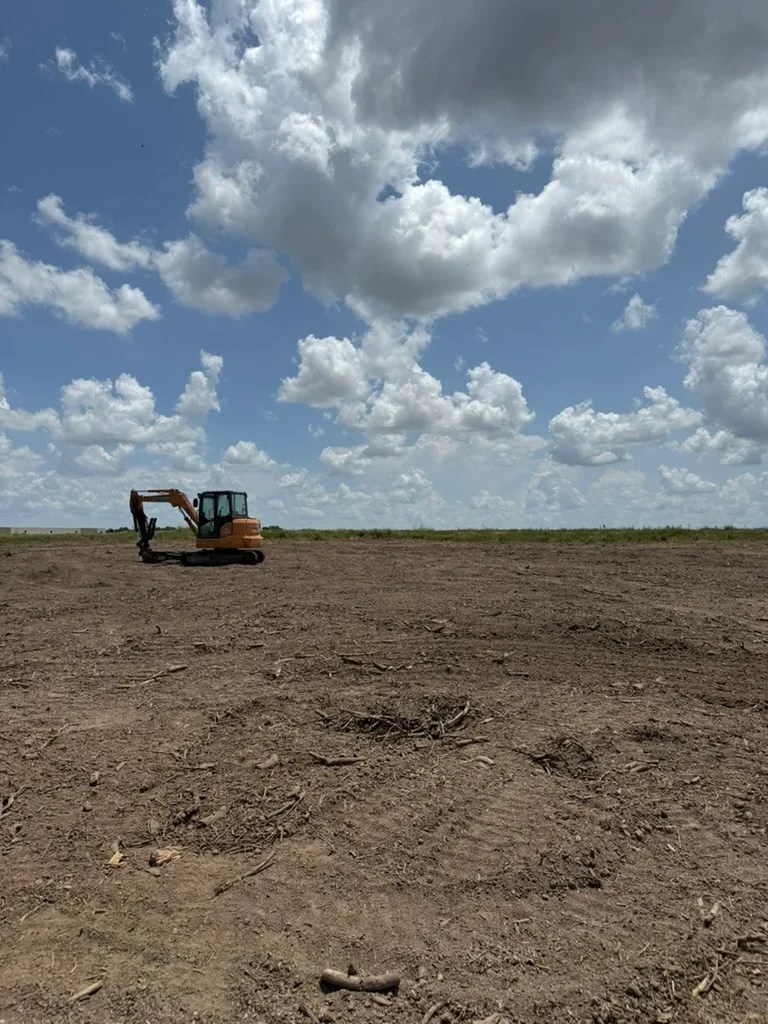Excavator clearing and grading open land with removed vegetation and debris visible