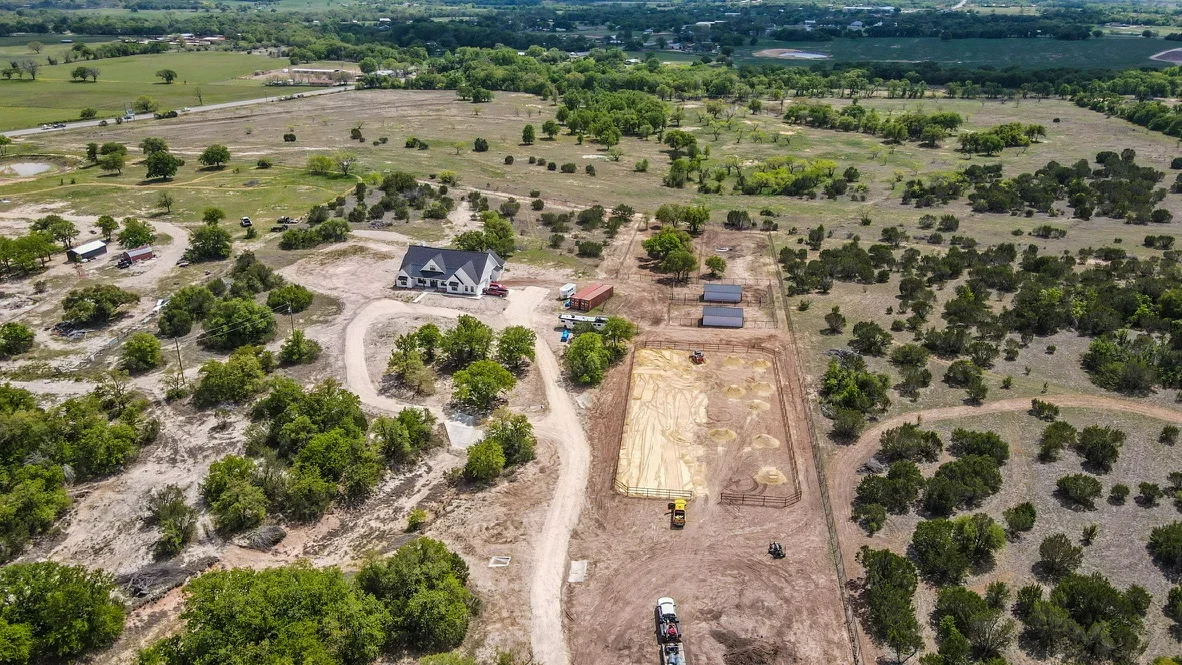 Aerial view of excavated residential site with curved driveway, graded foundation area, and preserved trees on rural acreage