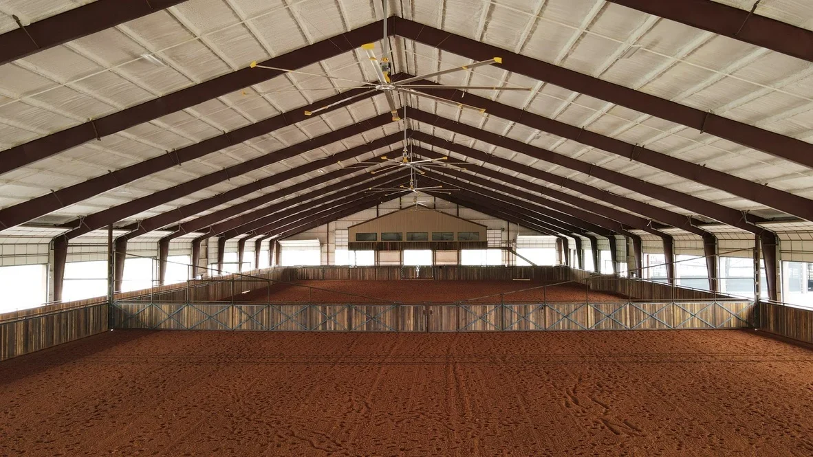 Indoor equestrian arena with steel frame, red clay footing, and wooden railings