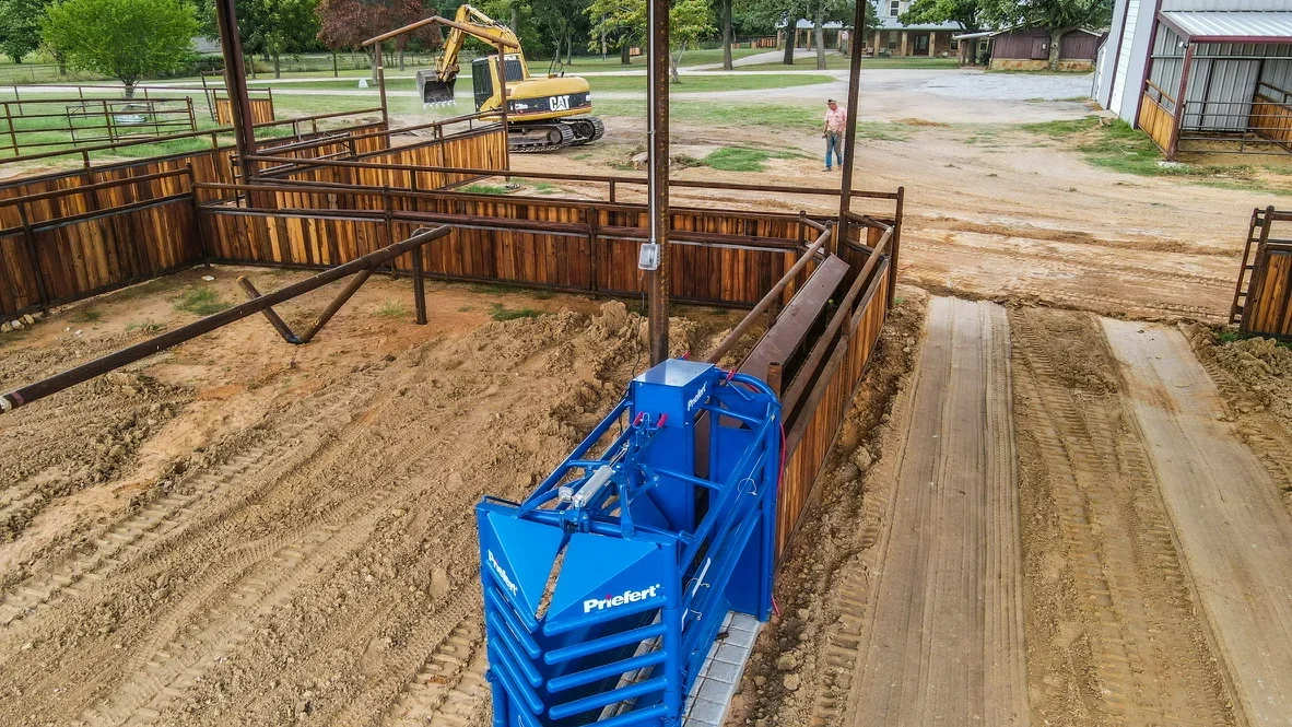 Excavated arena site with blue Prielert equipment, wooden fence panels, and graded soil base