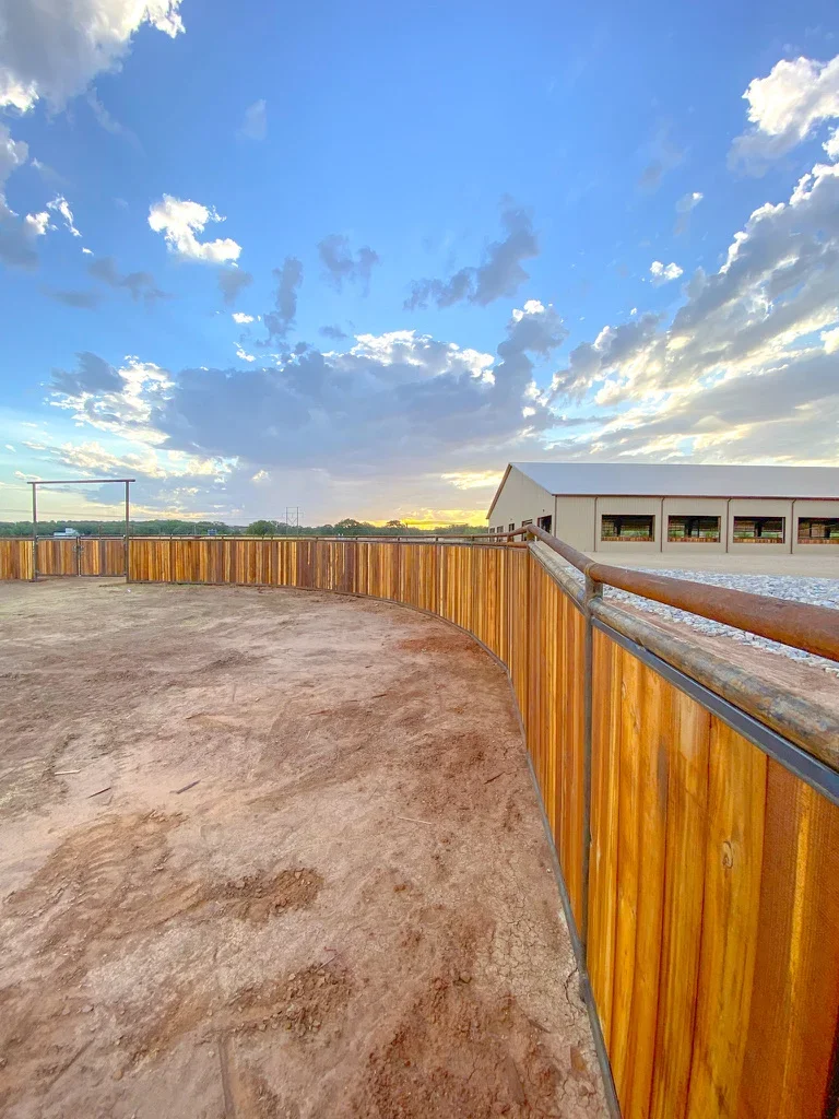 Graded dirt arena with wooden perimeter fence and metal rails, covered barn structure with stalls