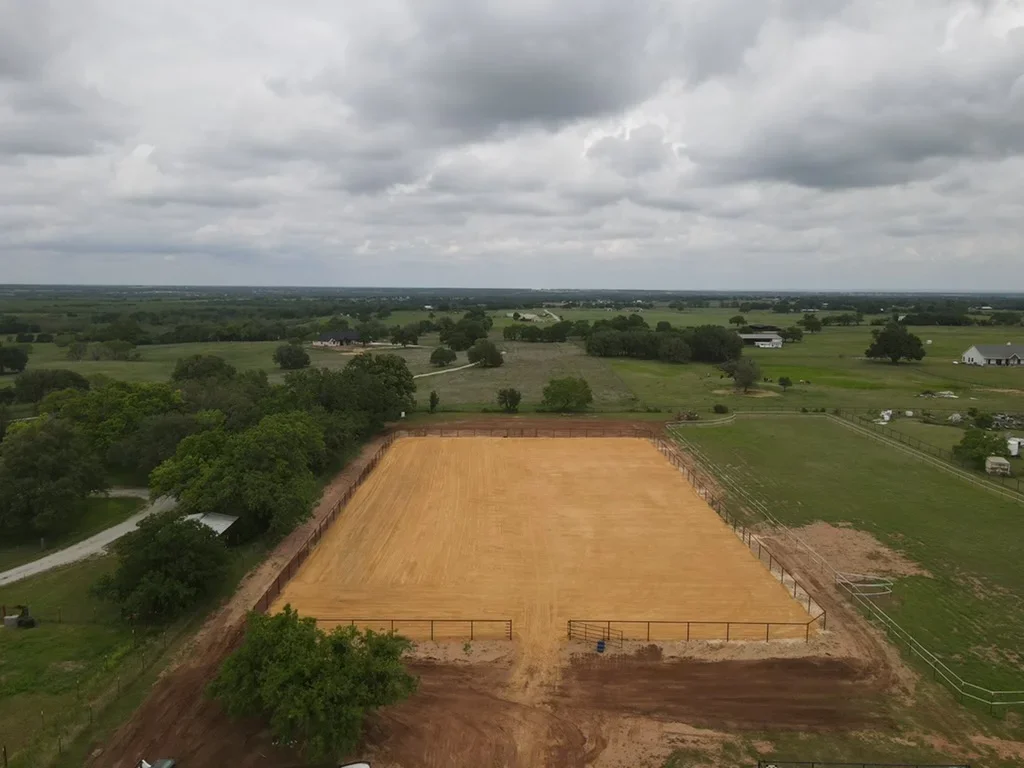 Aerial view of completed equestrian arena with clay surface, metal gates, and perimeter fencing