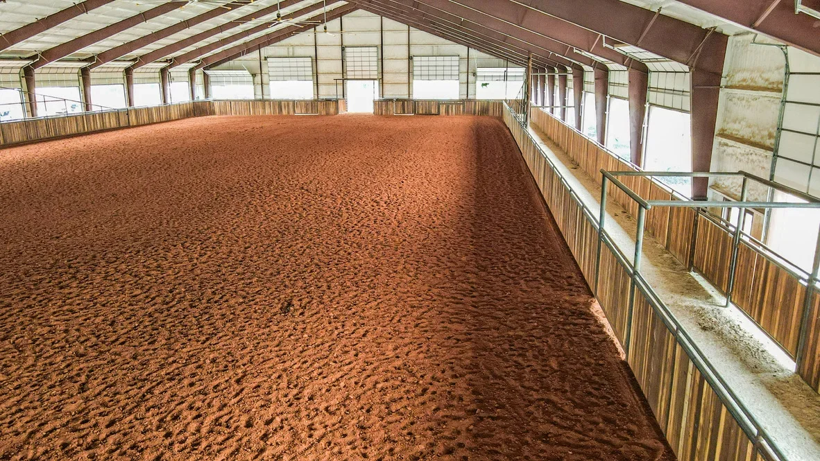 Compacted reddish-brown arena floor with wooden fencing and steel-frame structure with translucent roof panels