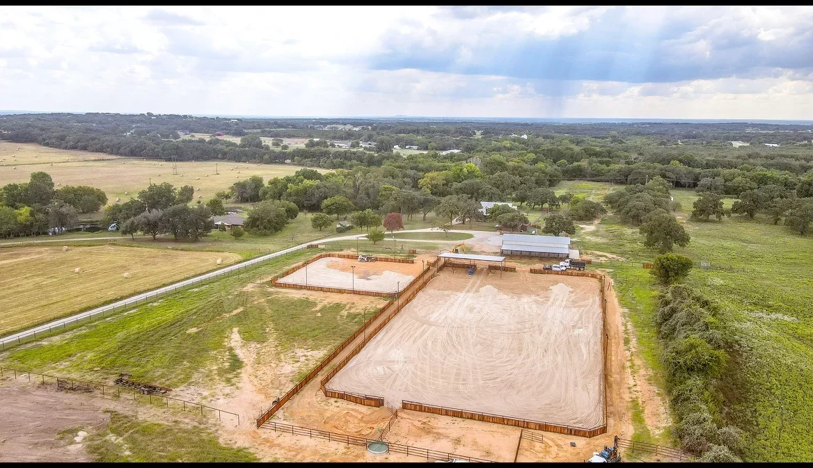 Aerial view of excavated equestrian arena with sand surface, wooden and metal fencing, and agricultural buildings