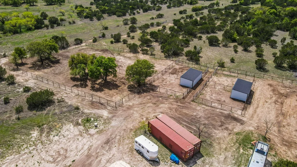 Aerial view of ranch with fenced pastures, metal pipe corrals, and blue shelter buildings on graded dirt