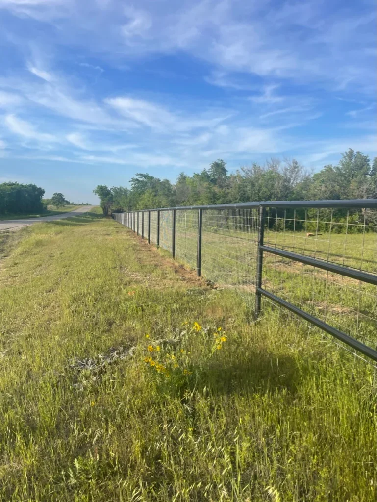 Steel pipe and wire mesh ranch fence installation along pasture with trees in background