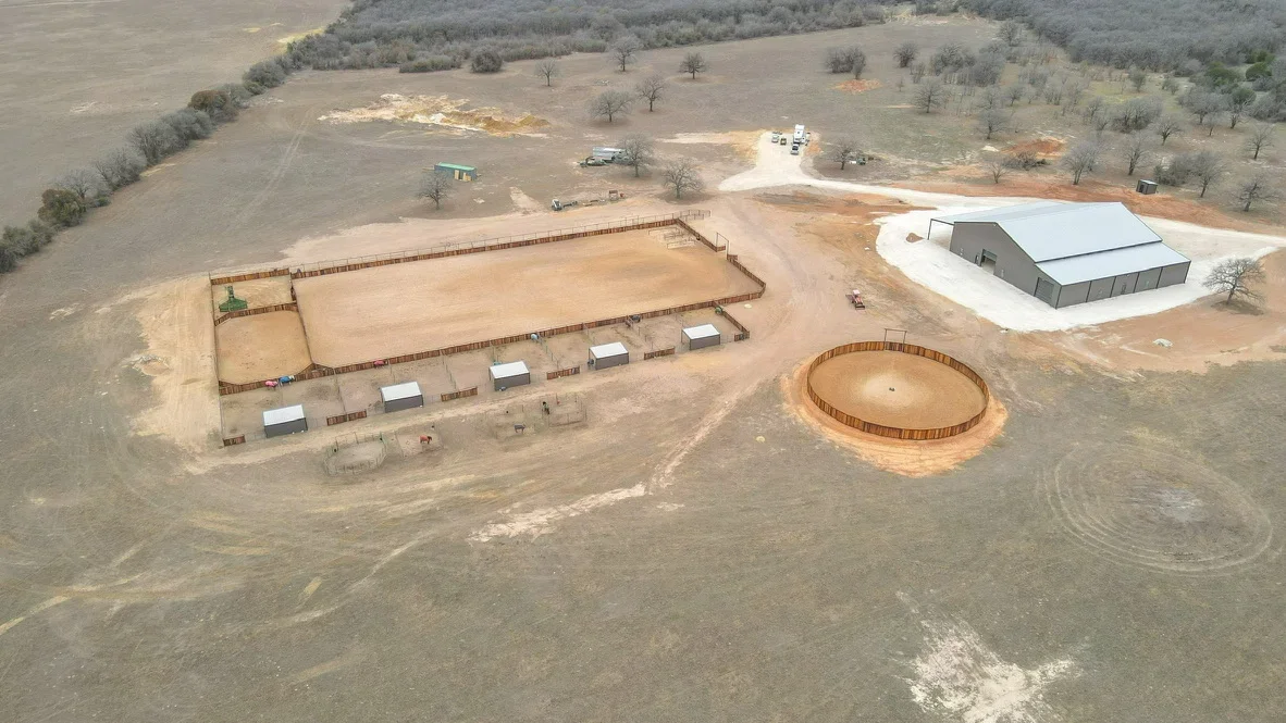 Aerial view of excavated equestrian facility with multiple arenas, wooden fencing, and metal structures on cleared land