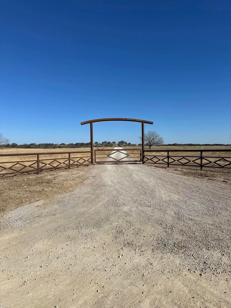 Wrought iron arched entry gate with diamond-pattern ranch fencing on gravel driveway