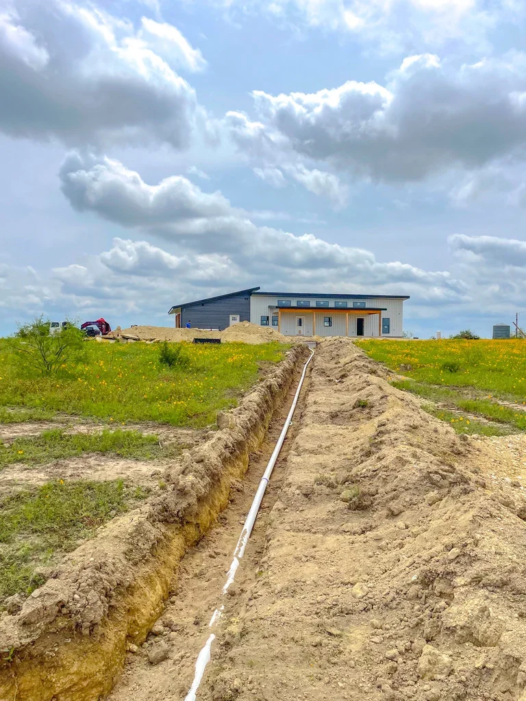 Excavated utility trench with white PVC pipe installed in prepared soil leading to residential building