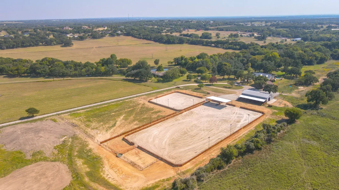 Aerial view of two prepared equestrian arenas with compacted sand base, metal fencing, and support buildings on rural property