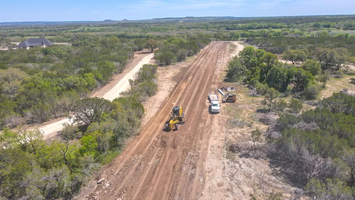 Motor grader working on dirt driveway with cleared shoulders and native soil base in rural setting