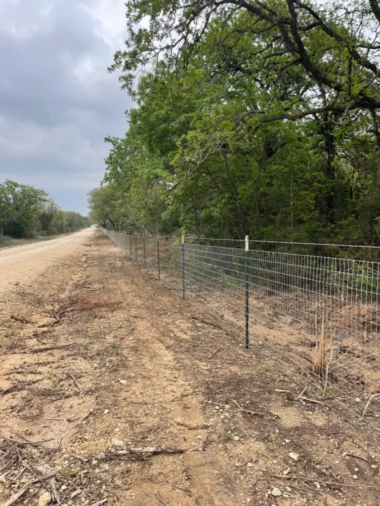 Welded wire fence with metal posts along graded dirt road bordered by mature trees