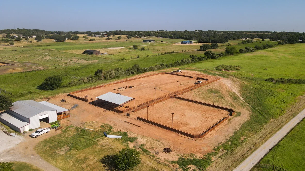 Aerial view of excavated equestrian arena with metal fencing, compacted red clay base, and metal-frame shelter structure