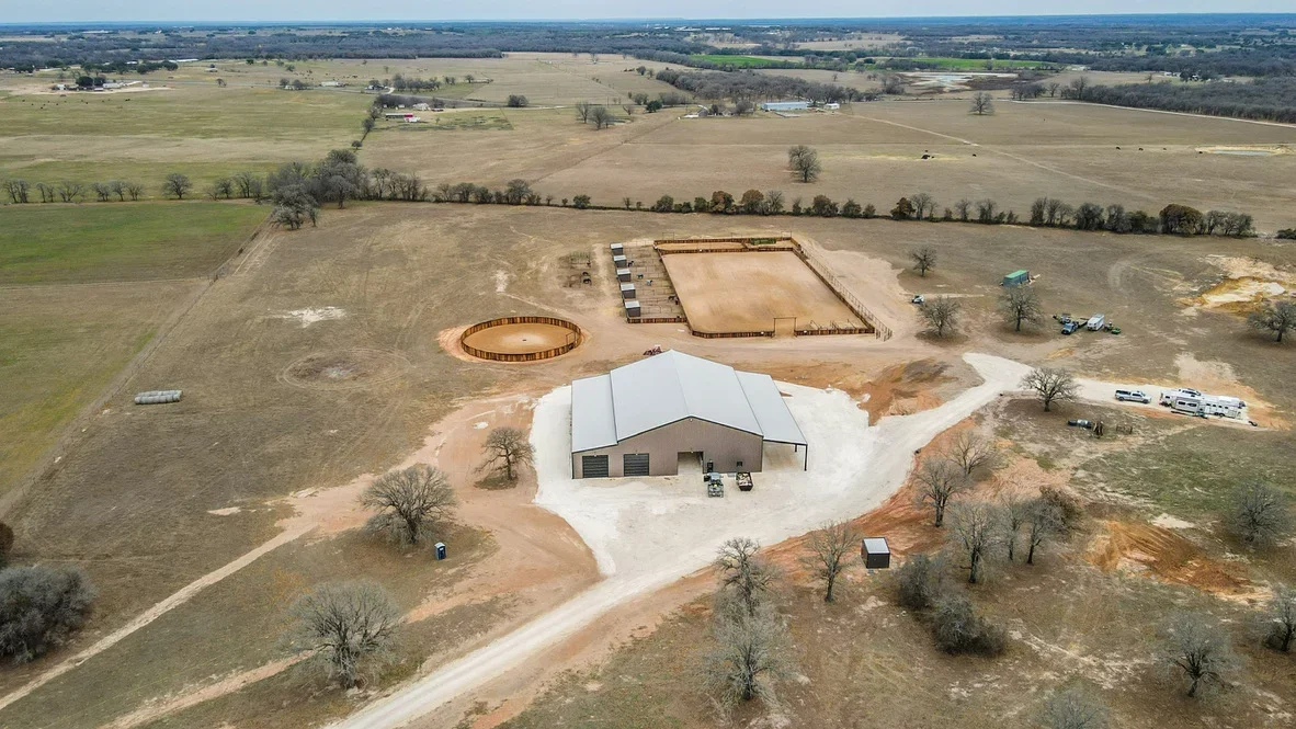 Aerial view of equestrian facility with metal-roofed arena, concrete pad, round pen, and fenced paddocks on cleared land