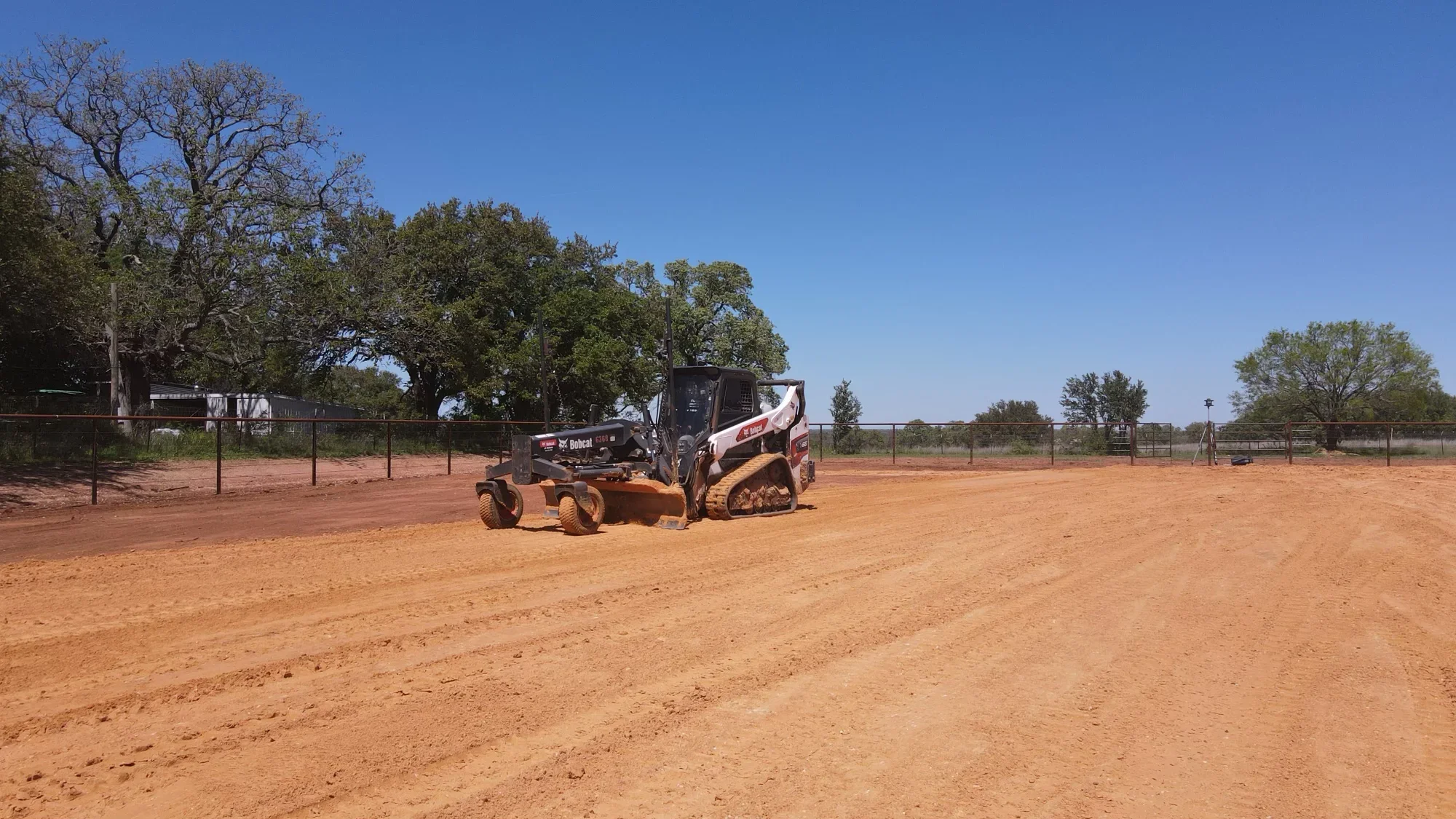 Bobcat track loader grading dirt arena with perimeter fencing and oak trees
