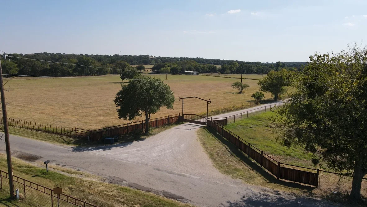 Asphalt driveway with metal archway gate and wooden ranch fencing on rural property