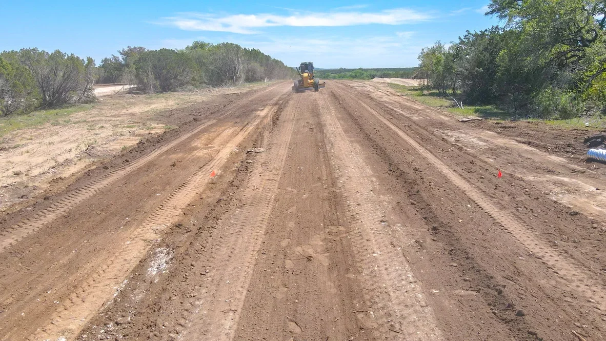 Motor grader working on unpaved road with graded dirt surface and cleared vegetation on both sides