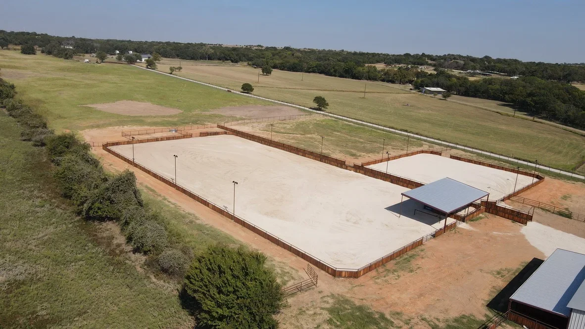 Aerial view of excavated recreational courts with concrete pads, metal fencing, and shelter structures on rural property