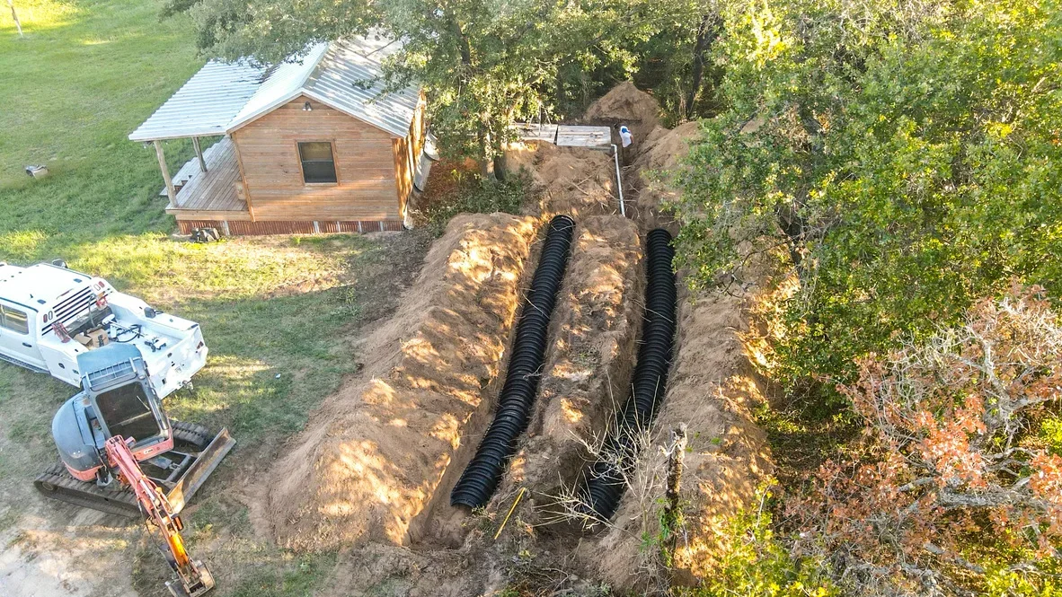 Aerial view of septic system installation with two black corrugated pipes in excavated trench near residential cabin