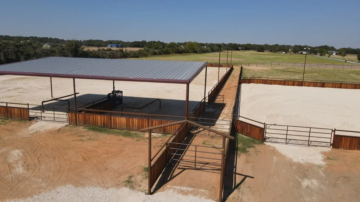 Equestrian facility with sand arenas, metal roof structure, steel fencing, and graded grounds