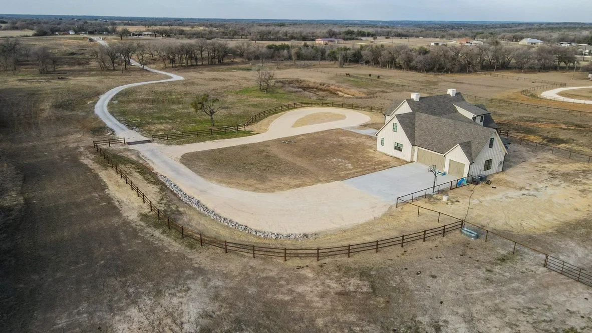 Aerial view of concrete driveway, arena pad, and curved access road with metal fencing on rural property