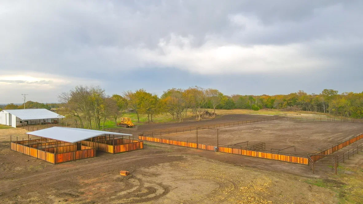 Excavated equestrian arena with metal fencing, shelter structures, and graded pasture areas