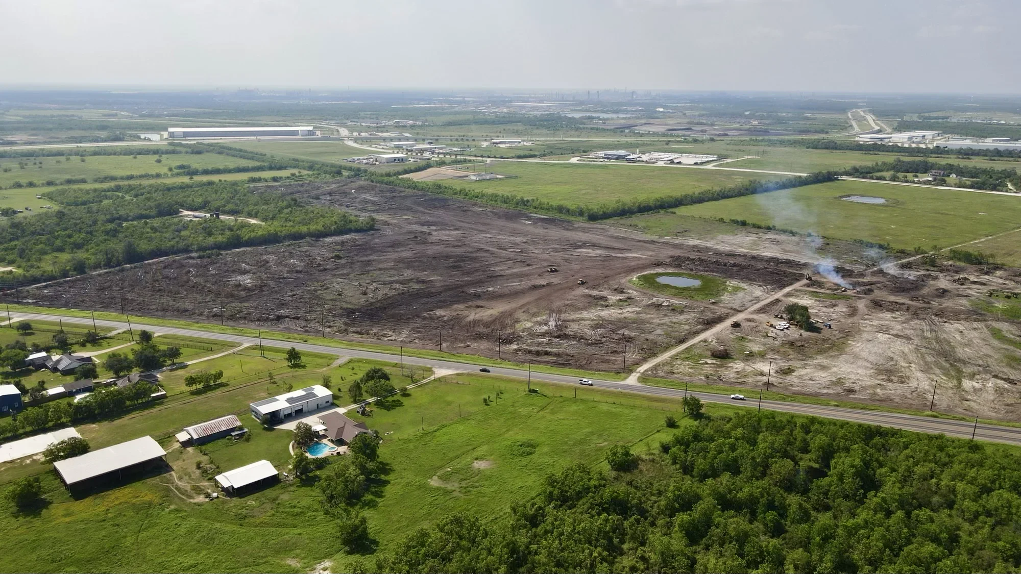 Aerial view of large cleared excavation site with exposed dark soil, heavy equipment, and surrounding green pasture