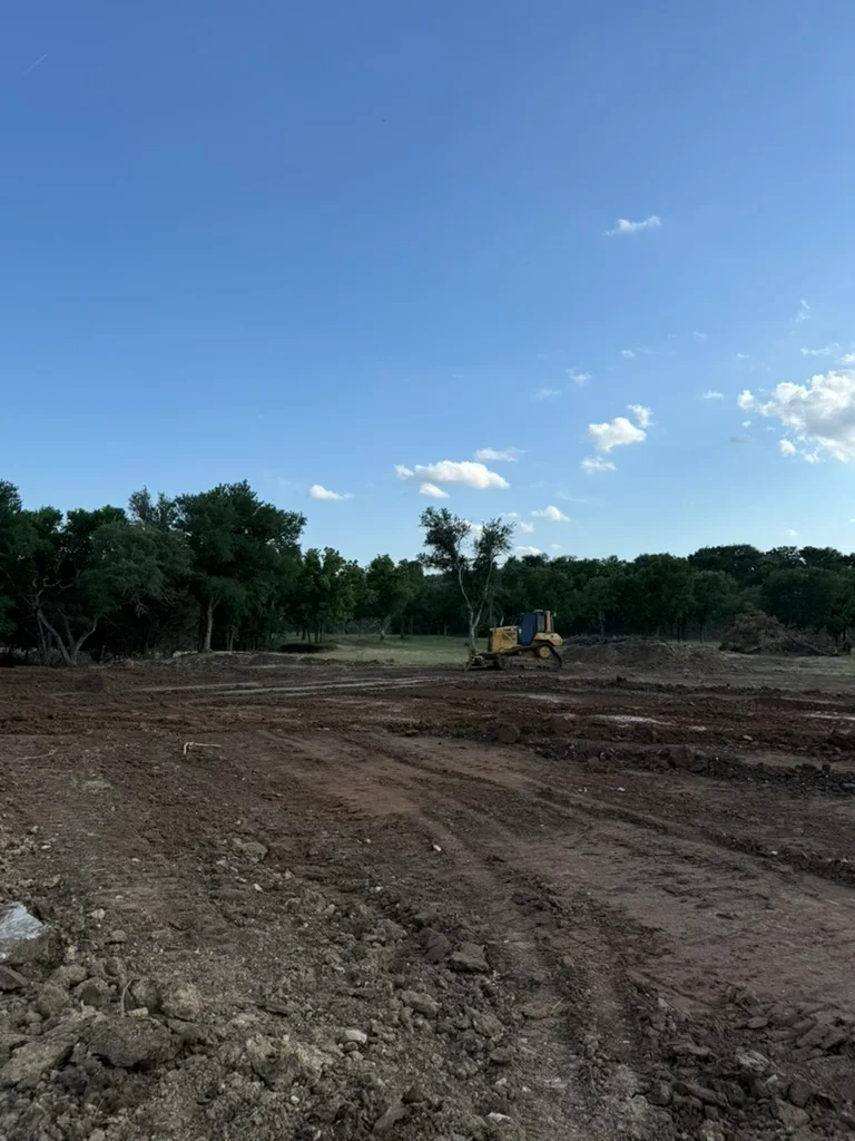 Cleared dirt lot with dozer, mature trees along edges, prepared building site