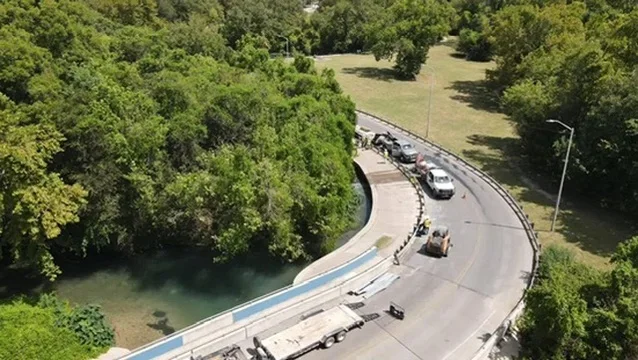 Curved roadway with blue erosion control matting, heavy equipment, and river embankment stabilization