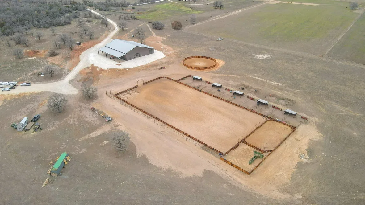 Aerial view of equestrian facility with two arenas, metal fencing, and agricultural building on prepared dirt site