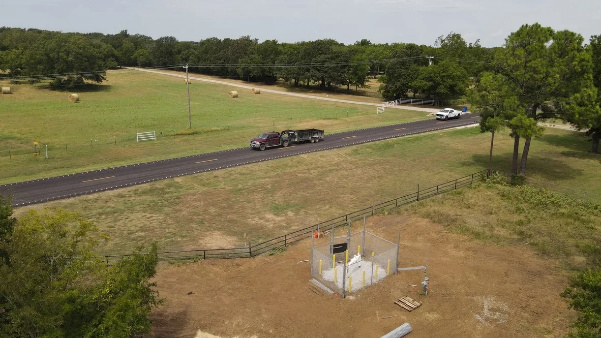 Asphalt driveway with white markings, metal ranch fencing, and graded earthwork on rural property